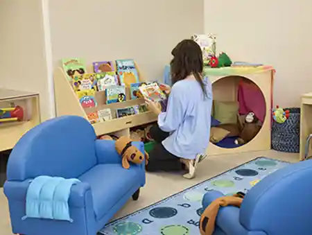 Woman in preschool classroom kneeling in front of bookshelf, looking at book